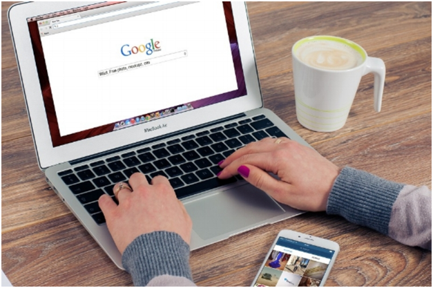 Person typing on a MacBook Air laptop with Google homepage open; coffee mug and smartphone are on the desk.