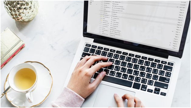 Person typing on a laptop with an email inbox open, next to a cup of tea and a notebook on a marble surface.